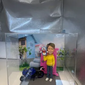 一個人像公仔的展示，公仔穿著黃色外套，手持一個叮噹（多啦夢）公仔，站在一輛藍色摩托車旁。背景是一座現代化的房子與綠意盎然的花園，整個展示在透明的展示盒中。