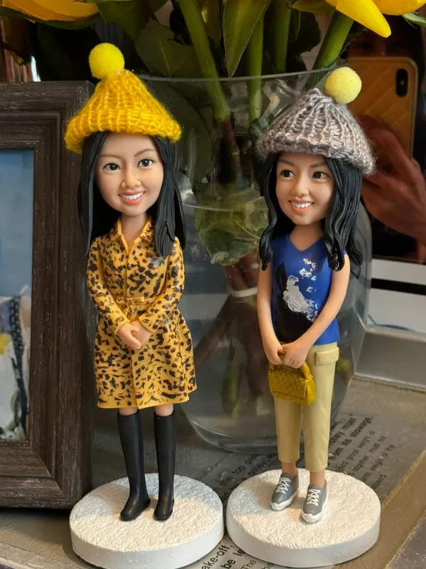 兩個手工製作的人像公仔，細緻還原女生的五官與穿搭，其中一位穿著黃色豹紋洋裝搭配黑色長靴，另一位穿著藍色上衣與淺色長褲，手持小手袋。兩個公仔都戴著可愛的毛線帽，非常適合作為聖誕節禮物或特別紀念品。