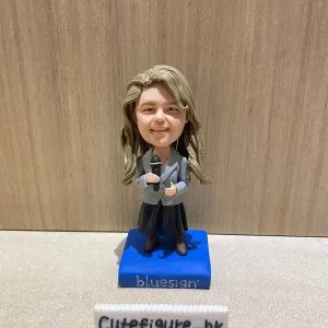 新聞發佈會主持人主題人像公仔：長捲髮女性身穿灰色西裝外套、藍色領帶與深色裙裝，雙手持黑色麥克風站在藍色方形底座上，底座印有 bluesign 標誌；前方擺放寫有 Cutefigure_hk 的小名牌，背景為木紋牆面。整體呈現正式發布會主持與新聞播報的職業形象，適合作為人像公仔榮休禮物與展示擺設。