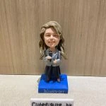 一位金髮女士的專業演講者人像公仔，她身穿灰色西裝外套，手持咪高峰，並站立在一個印有 "bluesign" 公司標誌的藍色地台上。