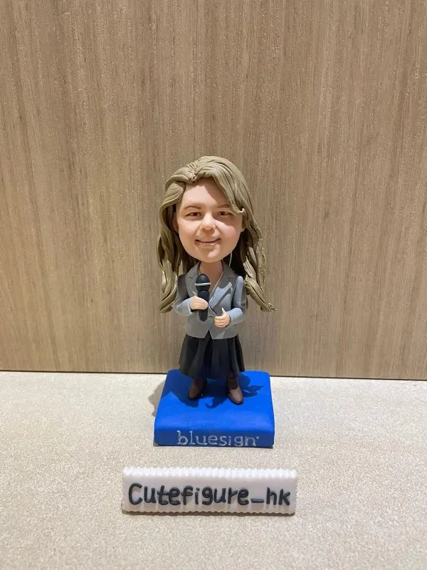 一位金髮女士的專業演講者人像公仔，她身穿灰色西裝外套，手持咪高峰，並站立在一個印有 "bluesign" 公司標誌的藍色地台上。