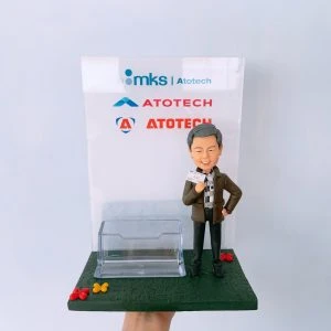 一座結合名片卡片座的商務風人像公仔擺設：灰髮微笑男子穿深色外套、格紋襯衫與黑色長褲，右手持名片、左手叉腰，站在深綠色長方形底座上；底座前緣點綴紅、黃小花。中前方配置透明塑料卡片座，可放置名片或卡片。背板為透明壓克力板，上方印有「mks | Atotech」與多個「ATOTECH」字樣與圖形標記。整體呈現乾淨俐落的辦公室桌面收納與展示效果，具紀念與實用功能。