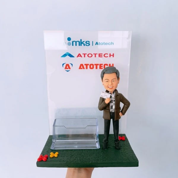 一座結合名片卡片座的商務風人像公仔擺設：灰髮微笑男子穿深色外套、格紋襯衫與黑色長褲，右手持名片、左手叉腰，站在深綠色長方形底座上；底座前緣點綴紅、黃小花。中前方配置透明塑料卡片座，可放置名片或卡片。背板為透明壓克力板，上方印有「mks | Atotech」與多個「ATOTECH」字樣與圖形標記。整體呈現乾淨俐落的辦公室桌面收納與展示效果，具紀念與實用功能。