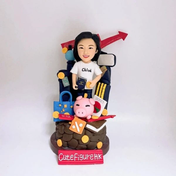 一座以理財與旅遊為主題的人像公仔：黑髮女子穿白色短袖T恤與黑裙，手持深藍色護照與金幣，站在滿載物件的場景前。周圍擺有藍色手提包、信用卡、粉紅小豬撲滿、金幣、紅色百分比折扣牌、書本、收據與計算器意象；背景有黑色行李箱、柱狀與箭頭上升的圖表，以及手機螢幕與鉛筆組，象徵投資成長與工作效率。整體以紅、藍、金為主色，底座寫著品牌字樣，呈現活潑俏皮的財務規劃與旅行熱愛主題。