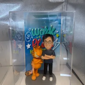 一個人像公仔和加菲貓的塑像展示在透明展示盒中。公仔穿著藍色T恤和黑色褲子，手臂交叉，旁邊的加菲貓站在灰色底座上，也同樣交叉手臂。背景有色彩繽紛的“Go Work”字樣，並搭配星星和閃電的裝飾，營造活潑氛圍。