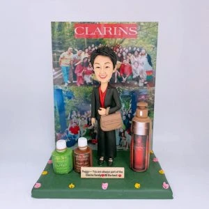 一款以告別與品牌致敬為主題的人像公仔:短髮女性身穿黑色套裝與紅色內搭,肩背米色側背包,站在綠色草地底座上。前方有刻字小牌寫上祝福語,左右擺放三款化妝品造型瓶罐;背景立牌為團體合照與品牌紅色標誌。整體呈現同事餞別與感謝主題,適合作為賤别禮物與人像公仔收藏。