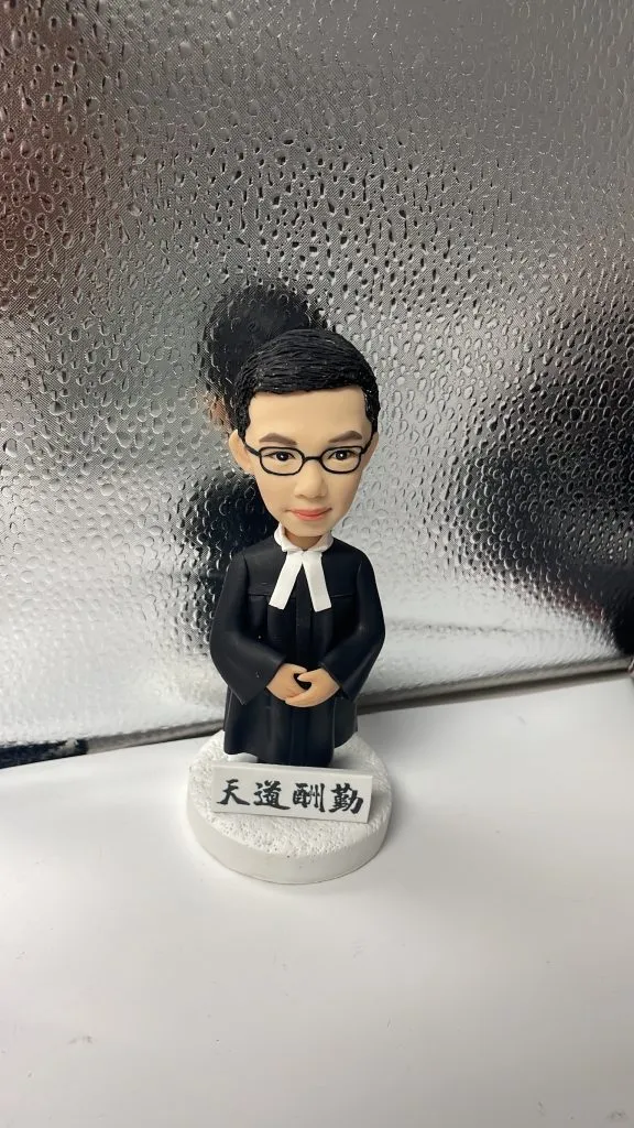 一款男性律師人像公仔，身穿律師袍，站立在刻有「天道酬勤」銘牌的底座上，是送給律師的完美入職禮物。
