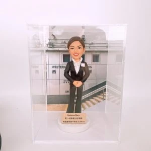 透明展示盒內，一尊女性警務主題的人像公仔端正站立：深色兩件式西裝外套配長褲，內搭白色荷葉邊襯衫，胸前別有識別證樣式的小牌；髮型為整齊盤髮，雙手於腹前交握。公仔立於白色圓形底座，底座前附金色銘牌，銘牌上可見英文名字與中文祝賀語，並提及「西區警署」及年份（2022）。展示盒背板為警署外牆與階梯的照片，牆面印有「WESTERN…」與中文「警署」字樣。整體色調乾淨，呈現升職賀禮的正式氛圍。