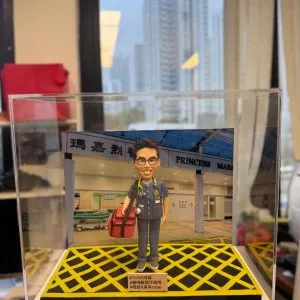 訂製醫生人像公仔｜醫護人員穿著深藍色醫護制服，胸前掛著聽診器，手持紅色醫療急救袋，背景為香港「瑪嘉烈醫院 Princess Margaret Hospital」正門場景。此公仔以急症室醫生為原型，作為專屬醫護禮物與紀念擺設。