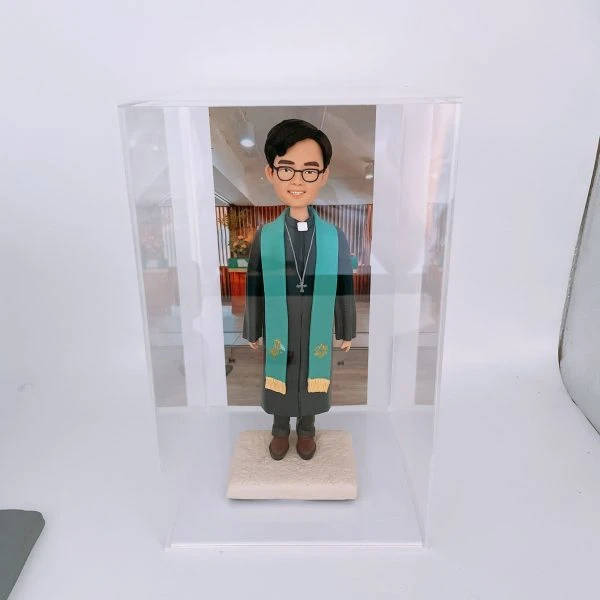 一款放置在透明亞加力膠盒中的客製化牧師人像公仔，牧師身穿聖袍，背後是教會照片背景，是一份莊重的感謝禮物。