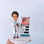 訂製醫生人像公仔｜身穿白袍的醫生公仔手持內視鏡器械，站在配有內視鏡影像顯示器與手術設備的機台旁，底座印有「Dr. Derek Fung」名字標牌。此醫護公仔細緻還原專業醫生形象，是專屬醫療禮物與紀念收藏。