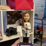 手掌托起的記者主題人像公仔：長髮女記者穿白色外套與黑色長褲，右手持麥克風、左手拿稿件，底座為深紫色圓形舞台並點綴白色燈泡，前方附有日期與字樣的小銘牌；背景為辦公室書架與攝影器材。整體呈現專業而溫暖的致敬氛圍，適合作為記者人像公仔榮休禮物與收藏擺設。