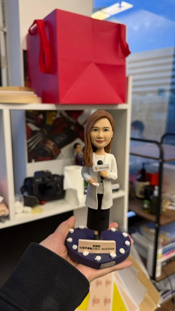 手掌托起的記者主題人像公仔：長髮女記者穿白色外套與黑色長褲，右手持麥克風、左手拿稿件，底座為深紫色圓形舞台並點綴白色燈泡，前方附有日期與字樣的小銘牌；背景為辦公室書架與攝影器材。整體呈現專業而溫暖的致敬氛圍，適合作為記者人像公仔榮休禮物與收藏擺設。