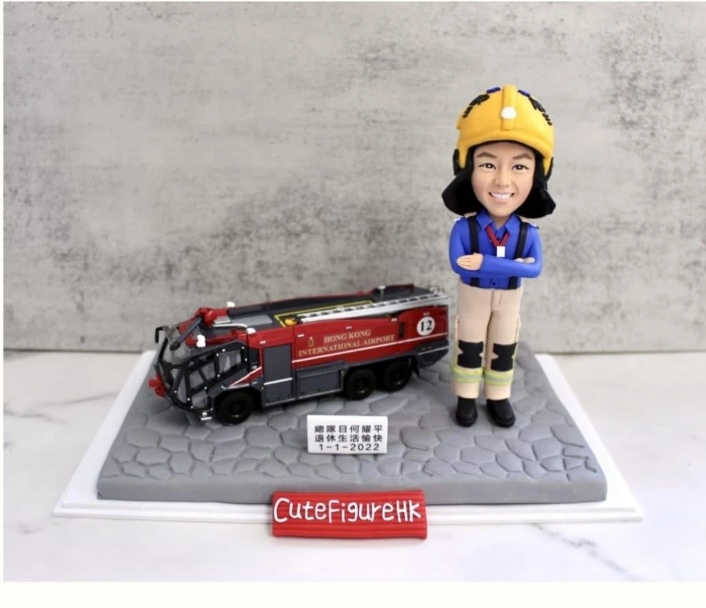 一款為機場消防員訂製的退休人像公仔，公仔身穿全套消防制服，旁邊停放著一輛香港國際機場的專用消防車模型，底座銘牌刻有贈予總隊目何耀平的退休祝福。