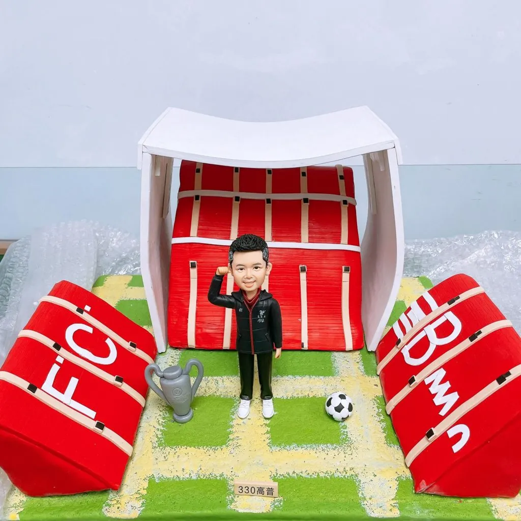 足球教練主題人像公仔：短髮男子穿黑色運動外套與長褲、白色球鞋，站在綠色方格草皮的足球場景上，右手舉臂加油；身旁有銀色冠軍獎盃與黑白足球。背景為紅白相間的看台與白色頂棚，兩側斜倒的紅色看台塊上可見字母裝飾，前方貼有小銘牌。整體呈現球場致敬與奪冠慶祝氛圍，適合作為足球教練的答謝禮物人像公仔。