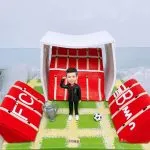 足球教練主題人像公仔：短髮男子穿黑色運動外套與長褲、白色球鞋，站在綠色方格草皮的足球場景上，右手舉臂加油；身旁有銀色冠軍獎盃與黑白足球。背景為紅白相間的看台與白色頂棚，兩側斜倒的紅色看台塊上可見字母裝飾，前方貼有小銘牌。整體呈現球場致敬與奪冠慶祝氛圍，適合作為足球教練的答謝禮物人像公仔。