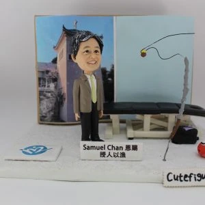 一款以「授人以漁」為主題的人像公仔：短髮男子穿西裝外套、淺綠色領帶與黑長褲，站在白色地台上，前方放有名牌，上寫「Samuel Chan 恩賜 授人以漁」。背景板左側為建築物照片，右側為淡藍色面板，上有黑色釣線示意；場景中擺放一張黑色治療/按摩床與工具箱，右前方立有灰色釣竿與紅色小魚模型；地台前緣有「Cutefigure_hk」字樣。整體呈教育與分享精神的釣魚人像公仔與人像公仔。