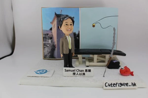 一款以「授人以漁」為主題的人像公仔：短髮男子穿西裝外套、淺綠色領帶與黑長褲，站在白色地台上，前方放有名牌，上寫「Samuel Chan 恩賜 授人以漁」。背景板左側為建築物照片，右側為淡藍色面板，上有黑色釣線示意；場景中擺放一張黑色治療/按摩床與工具箱，右前方立有灰色釣竿與紅色小魚模型；地台前緣有「Cutefigure_hk」字樣。整體呈教育與分享精神的釣魚人像公仔與人像公仔。