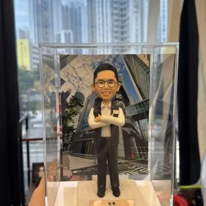 一尊以警務處「警長」職級為主題的人像公仔置於透明壓克力展示盒中，由左手掌托起近距離拍攝。公仔五官細緻：短而整齊的黑髮、劉海向上梳理；配戴圓框眼鏡，露出齒列微笑。上身著白色立領襯衫，袖口整潔收邊；外搭黑色無袖戰術背心，面料呈細霧面質感。左胸配有長條形識別牌與織章貼片，右胸口袋蓋上有淺色方形證章飾片與小型徽記；背心立體口袋、門襟與下擺車縫線條清晰。下身為深灰長褲，褲線筆直，腳穿黑色皮鞋，鞋面有亮面反光。公仔雙臂在胸前自然交疊，手掌比例圓潤，呈沉著自信的站姿。腳下為米白色粗紋理台座，前緣安置金色金屬名牌，刻有「祝賀…警長 榮陞之喜」等字樣（其餘字樣略）。展示盒後方插入高樓林立的商業街景背景板，仰角視角可見玻璃帷幕反射雲影與樹梢。更外層透過辦公室落地窗可見實景住宅高樓群與藍天，窗面映出長條燈具反光；畫面左側下方可見毛絨外套袖口與桌面書籍、文具與收納盒等物件，強化在職場環境中呈交禮的情境。