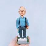一款為外國老闆訂製的退休禮物人像公仔，公仔是一位戴眼鏡的年長男士，身穿藍色襯衫和吊帶褲，站立在一個刻有「Happy Retirement」祝福語的石板路底座上。