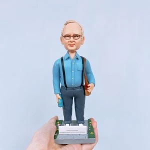 一款為外國老闆訂製的退休禮物人像公仔，公仔是一位戴眼鏡的年長男士，身穿藍色襯衫和吊帶褲，站立在一個刻有「Happy Retirement」祝福語的石板路底座上。