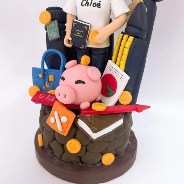 一座以理財與旅遊為主題的人像公仔：黑髮女子穿白色短袖T恤與黑裙，手持深藍色護照與金幣，站在滿載物件的場景前。周圍擺有藍色手提包、信用卡、粉紅小豬撲滿、金幣、紅色百分比折扣牌、書本、收據與計算器意象；背景有黑色行李箱、柱狀與箭頭上升的圖表，以及手機螢幕與鉛筆組，象徵投資成長與工作效率。整體以紅、藍、金為主色，底座寫著品牌字樣，呈現活潑俏皮的財務規劃與旅行熱愛主題。