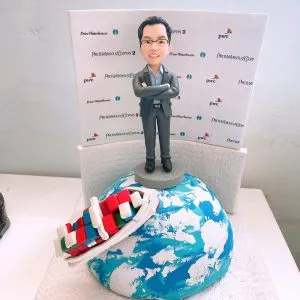 短髮戴眼鏡的男士穿灰色西裝與淺藍襯衫,雙手抱胸站在灰色小圓台上;腳下為藍白相間的地球造型底座,左側斜倚一艘以紅、綠、藍、白方塊色塊呈現的郵輪模型。背板印有多個公司字樣與標誌圖案。整體置於白色紋理平台上,風格專業又具旅遊度假意象,適合作為老闆退休的專屬人像公仔退休禮物。