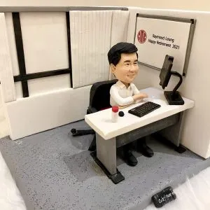 一座辦公室情境的人像公仔：短髮戴眼鏡的男子穿白襯衫與黑皮鞋，坐在黑色辦公椅上，雙臂自然靠在白色長方形辦公桌。桌上擺有單螢幕顯示器（黑色可調臂座）、長條鍵盤、滑鼠與一枚紅白公章。右後方白色看板印有圓形紅色標記與「Raymond Leung Happy Retirement 2021」致意字樣；周圍以白色隔板與灰色地台營造開放式隔間辦公室的氛圍。整體呈現簡潔沉穩的職場與榮退紀念場景。