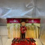 透明展示盒內的一款定製人像公仔：背景為紅底金字「××酒家」拱門與垂幕，象徵中式酒樓／餐館；公仔塑成中年女店主形象，短髮、笑容親切，穿紅色繡花上衣配黑長褲與涼鞋，右手持麥克風作開幕致辭。腳下紅色小舞台點綴暖黃燈串，前方陳列多個黑色音響／麥克風座，整體呈現宴會式開張氛圍。台前示例字牌寫「生日快樂」，實際下單可改為店名與開業賀詞，適合作為店鋪開張、新店啟業的人像公仔禮物。