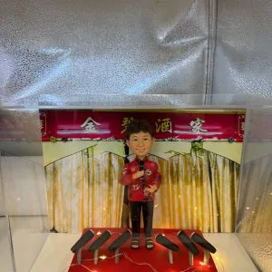 透明展示盒內的一款定製人像公仔：背景為紅底金字「××酒家」拱門與垂幕，象徵中式酒樓／餐館；公仔塑成中年女店主形象，短髮、笑容親切，穿紅色繡花上衣配黑長褲與涼鞋，右手持麥克風作開幕致辭。腳下紅色小舞台點綴暖黃燈串，前方陳列多個黑色音響／麥克風座，整體呈現宴會式開張氛圍。台前示例字牌寫「生日快樂」，實際下單可改為店名與開業賀詞，適合作為店鋪開張、新店啟業的人像公仔禮物。
