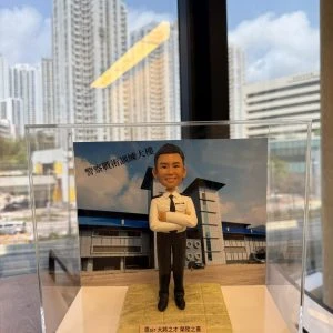 一款為香港警務人員訂製的晉升人像公仔，公仔身穿白色警官制服，背景為警察戰術訓練大樓，用以祝賀其榮陞之喜。