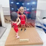 籃球場景雙人籃球人像公仔：一位身穿紅色23號球衣的籃球明星造型公仔站在籃框前微笑致意，另一位穿紅色45號球衣的年輕球迷公仔在前景持球仰頭準備投籃；木紋球場地台附有金色小銘牌寫著「Happy life begins…」，背景是燈光明亮的室內球館與籃板框，整體呈現偶像與粉絲在球場互動的溫馨畫面。