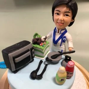 藍白配色廚師服的人像公仔站在圓形台座上微笑；頸掛藍色領結與胸針徽章，象徵專業證書。台面擺有小型烤箱、兩口平底鍋與黑色料理匙、綠白分層蛋糕、以及黃色與紅色調味瓶，呈現烘焙與西餐場景。整體精緻典雅，適合作為藍帶廚師考證祝賀禮物與收藏擺設。