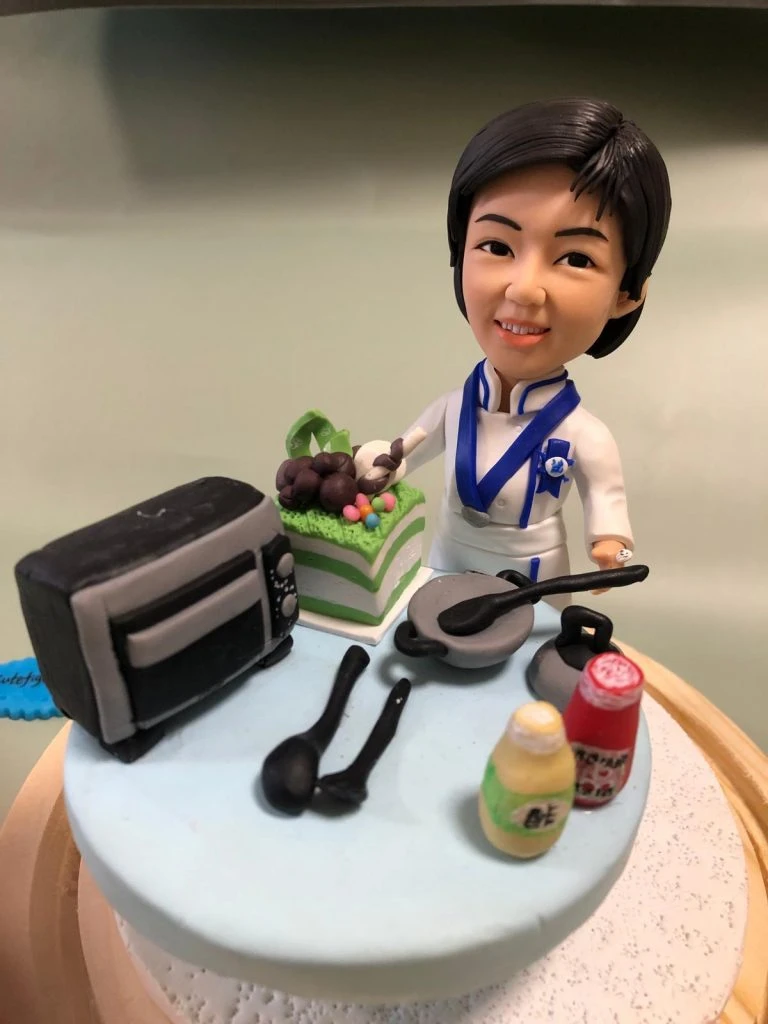 藍白配色廚師服的人像公仔站在圓形台座上微笑；頸掛藍色領結與胸針徽章，象徵專業證書。台面擺有小型烤箱、兩口平底鍋與黑色料理匙、綠白分層蛋糕、以及黃色與紅色調味瓶，呈現烘焙與西餐場景。整體精緻典雅，適合作為藍帶廚師考證祝賀禮物與收藏擺設。