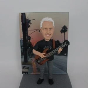 一位銀髮男士的樂手人像公仔,他身背一把低音結他,面帶微笑,站立在一塊以加州黃昏棕櫚樹街景為主題的相片背景板前。