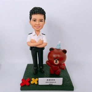 一款為香港紀律部隊人員訂製的榮休人像公仔，主角身穿白色制服，身旁站著一隻戴派對帽的Brown熊大卡通公仔作為背景配件裝飾。