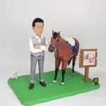 馬術主題人像公仔場景：短髮男子公仔身穿白襯衫搭灰色背心與長褲，雙手抱胸站在綠色草地底座上；身旁是一匹棕色馬，配有藍色韁繩與鞍具、白色鞍毯。右側豎立木質指示牌（附書法字樣），地面另有小型裝飾物。整體呈現馬場教學與照護氛圍，適合作為致敬馬術師的退休禮物人像公仔。
