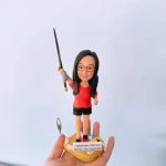 一款女性釣魚主題的人像公仔站在海灘風底座上：長直黑髮、紅色眼鏡，穿紅色上衣與黑色短褲、紅黑運動鞋。右臂高舉黑灰相間的釣竿，細釣線垂下，左側懸著小魚模型；底座有貝殼、救生圈等沙灘元素，前方貼有名牌「Dispenser, TSK VP Wong Yuen Yue Doris」。