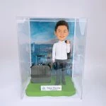 一款保險主題的人像公仔置於透明展示盒內：短髮男子微笑站立，穿白色襯衫、深色長褲與黑色皮鞋，右肩隨性搭著深色西裝外套；身旁放置一個深灰色拉桿登機箱。腳下為草綠色底座，前方小牌印有「Tokio Marine Hong Kong」與品牌標誌；背板是高樓林立、臨海的城市天際線照片，營造出差與專業形象。整體呈現勤勉專業、常出差拜訪客戶的保險人像公仔與人像公仔。