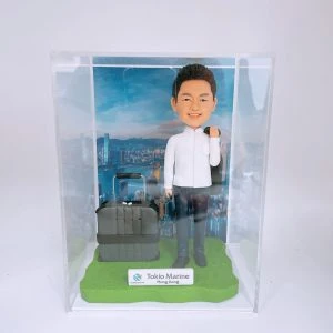 一款保險主題的人像公仔置於透明展示盒內:短髮男子微笑站立,穿白色襯衫、深色長褲與黑色皮鞋,右肩隨性搭著深色西裝外套;身旁放置一個深灰色拉桿登機箱。腳下為草綠色底座,前方小牌印有「Tokio Marine Hong Kong」與品牌標誌;背板是高樓林立、臨海的城市天際線照片,營造出差與專業形象。整體呈現勤勉專業、常出差拜訪客戶的保險人像公仔與人像公仔。