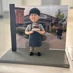 咖啡店主題人像公仔擺設：捲髮笑臉的店主穿淺灰上衣與深灰圍裙，雙手捧著巧克力分層蛋糕站在灰色質感底座上；左側有仿木製店招立牌，背板為咖啡店門面與綠植街景照片。整體呈現開業氣氛與手作風格，適合作為咖啡店開張禮物與店內展示擺設。