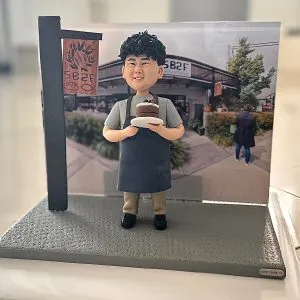 咖啡店主題人像公仔擺設：捲髮笑臉的店主穿淺灰上衣與深灰圍裙，雙手捧著巧克力分層蛋糕站在灰色質感底座上；左側有仿木製店招立牌，背板為咖啡店門面與綠植街景照片。整體呈現開業氣氛與手作風格，適合作為咖啡店開張禮物與店內展示擺設。