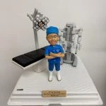 醫生人像公仔，身穿藍色手術服與口罩帽，雙手交叉站立，背景搭配手術床、手術燈及達文西手術機械臂模型。此公仔為醫生專屬客製化禮物，適合作為醫護紀念品、感謝醫生的特別禮物。