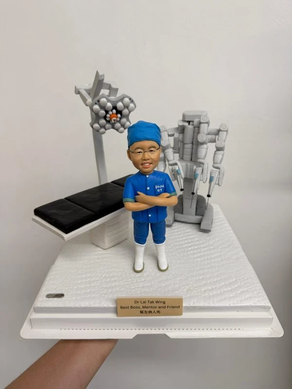 醫生人像公仔，身穿藍色手術服與口罩帽，雙手交叉站立，背景搭配手術床、手術燈及達文西手術機械臂模型。此公仔為醫生專屬客製化禮物，適合作為醫護紀念品、感謝醫生的特別禮物。