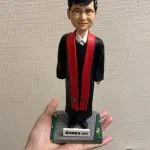 單人牧師人像公仔：黑色長袍外搭紅色聖帶，白色立領襯衫領口微露，配戴眼鏡、短黑髮，表情溫和端莊；立於灰色石板感底座，四角綠邊點綴小花，前方銘牌可客製年份與祝福文字。此人像公仔適合作為榮休、授職、周年與致謝的禮物。