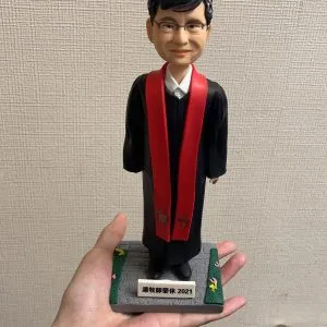單人牧師人像公仔：黑色長袍外搭紅色聖帶，白色立領襯衫領口微露，配戴眼鏡、短黑髮，表情溫和端莊；立於灰色石板感底座，四角綠邊點綴小花，前方銘牌可客製年份與祝福文字。此人像公仔適合作為榮休、授職、周年與致謝的禮物。