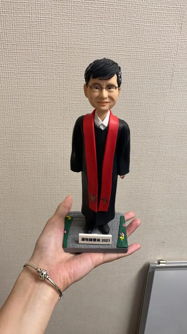 單人牧師人像公仔：黑色長袍外搭紅色聖帶，白色立領襯衫領口微露，配戴眼鏡、短黑髮，表情溫和端莊；立於灰色石板感底座，四角綠邊點綴小花，前方銘牌可客製年份與祝福文字。此人像公仔適合作為榮休、授職、周年與致謝的禮物。