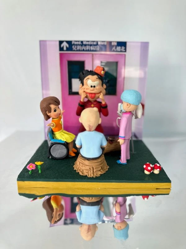 兒科病房場景中的醫生與醫護人像公仔：背景是醫院兒科病房入口（兒科病科病房 / Paed. Medical Ward），場景中有一位戴藍色手術帽、身穿粉紅制服的醫護公仔，陪伴兩位小病童──一位坐輪椅的小女孩、一位頭上有手術疤痕的男孩；中央則有一位扮小丑的表演者，用誇張表情逗笑病童。展示盒細節包含花朵、紅白蘑菇裝飾，呈現出溫馨療癒氛圍，適合作為醫生醫護主題的禮物或紀念公仔。