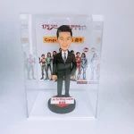 一款保險公司主題的人像公仔：短髮男子身穿黑色西裝與條紋領帶，微笑站在圓形底座上，前方名牌顯示英文與中文姓名。整體置於透明展示盒內，背板為公司活動視覺，醒目標示公司175周年與品牌標誌，並有身穿英雄風格戰服的團隊形象圖與日期標記。作品呈現專業穩重與慶典氛圍，適合保險團隊的里程碑紀念。
