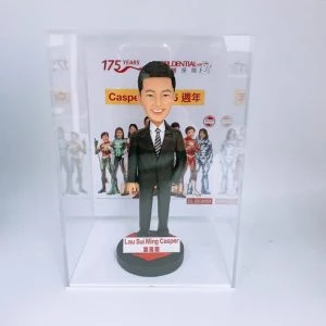 一款保險公司主題的人像公仔:短髮男子身穿黑色西裝與條紋領帶,微笑站在圓形底座上,前方名牌顯示英文與中文姓名。整體置於透明展示盒內,背板為公司活動視覺,醒目標示公司175周年與品牌標誌,並有身穿英雄風格戰服的團隊形象圖與日期標記。作品呈現專業穩重與慶典氛圍,適合保險團隊的里程碑紀念。